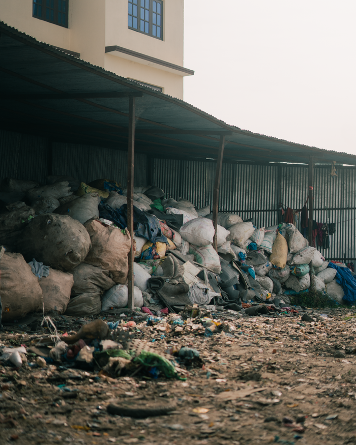 Piles of garbage bags stacked in an outdoor waste disposal area near industrial buildings.
